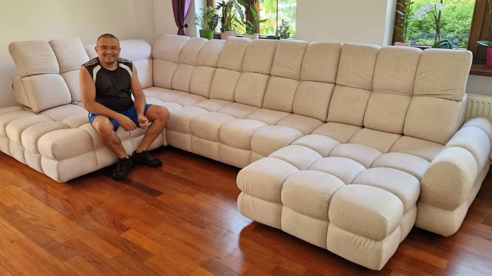 Man sitting on a beige Tollo XL u shape sofa in a room with wooden flooring and large windows.